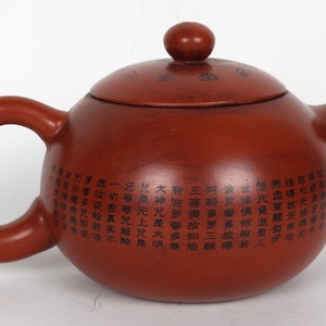 Peut inclure: Une théière en argile rouge avec un corps rond et un couvercle. La théière a une poignée et un bec verseur. La théière est décorée de caractères chinois noirs.