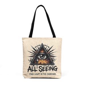 May include: A beige tote bag with black handles featuring a graphic of a triangle containing an eye, flames, and a sun. Below the graphic, the text reads ALL SEEING and FIND LIGHT IN THE SHADOWS in a black, dripping, stylized font.