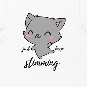 Op de afbeelding: Wit T-shirt met een cartoon grijze kat met roze wangen en de tekst "just keep stimming" in zwart.