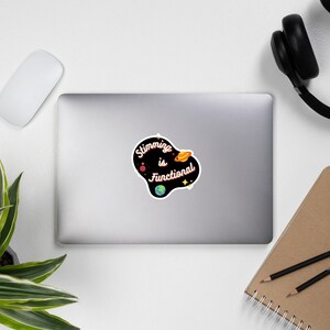 Peut inclure: Un autocollant noir et blanc avec un contour blanc. L'autocollant pr&eacute;sente un fond noir avec un motif de plan&egrave;te blanc, jaune et rouge. Le texte "Stimming is Functional" est &eacute;crit en blanc avec un contour rose.