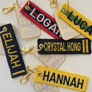 May include: A collection of personalized keychains with embroidered names in various colors. The keychains feature names like "Logan," "Lucas," "Elijah," "Crystal Hong," and "Hannah" in white or yellow thread. Each keychain has a gold-colored clasp.