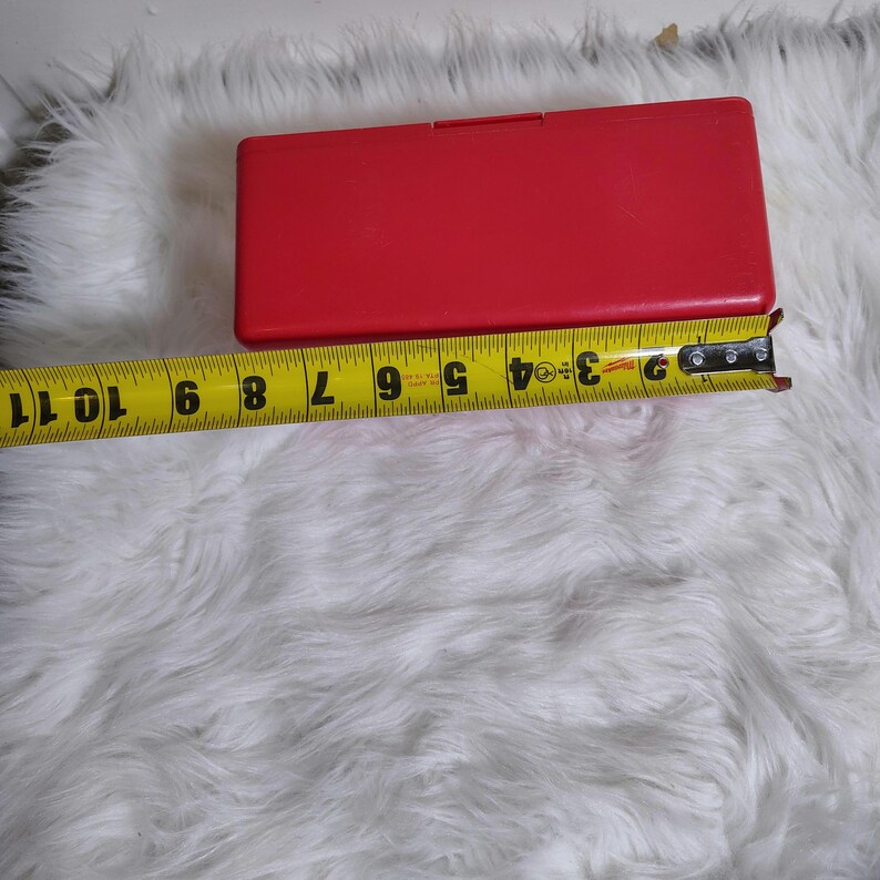 K&ouml;nnte beinhalten: Eine leuchtend rote rechteckige Box mit einem Klappdeckel, etwa 25 cm lang, wird gezeigt. Ein gelbes Ma&szlig;band liegt neben der Box und zeigt ihre L&auml;nge an. Die Box befindet sich auf einer wei&szlig;en, flauschigen Oberfl&auml;che.