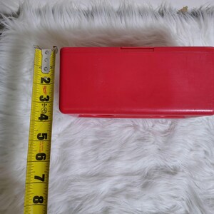 K&ouml;nnte beinhalten: Eine leuchtend rote, rechteckige Kunststoffbox mit einem Klappdeckel. Die Box ist etwa 18 cm lang. Ein gelbes Ma&szlig;band liegt neben der Box und zeigt die L&auml;nge in Zoll an.