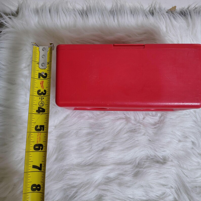 K&ouml;nnte beinhalten: Eine leuchtend rote, rechteckige Kunststoffbox mit einem Klappdeckel. Die Box ist etwa 18 cm lang. Ein gelbes Ma&szlig;band liegt neben der Box und zeigt die L&auml;nge in Zoll an.