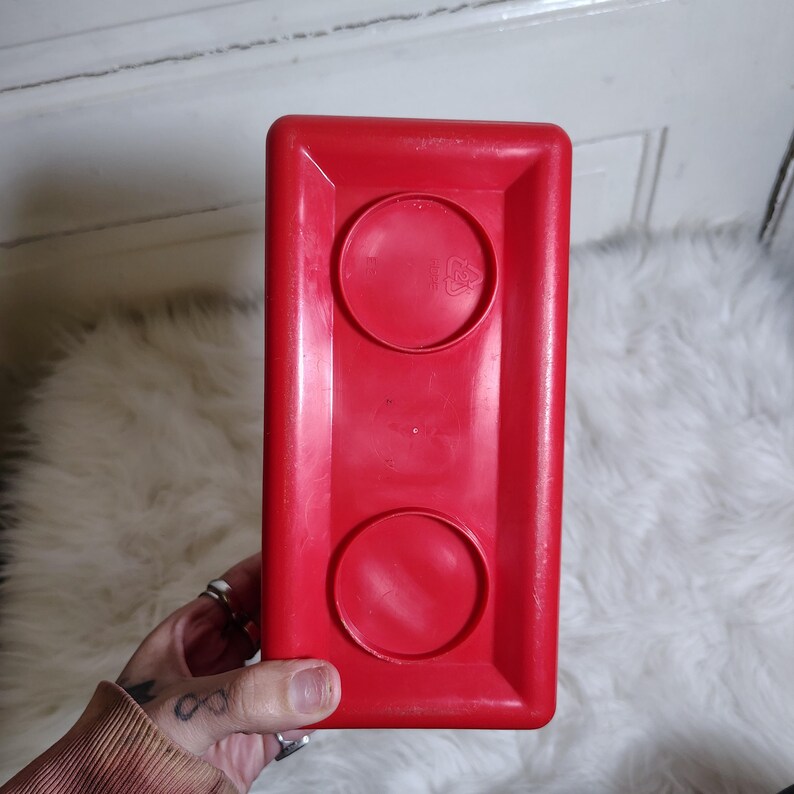 K&ouml;nnte beinhalten: Ein leuchtend roter Kunststoffbeh&auml;lter mit zwei kreisf&ouml;rmigen Vertiefungen. Der rechteckige Beh&auml;lter wird von einer Hand gehalten, vor einem wei&szlig;en, flauschigen Hintergrund. Der Beh&auml;lter hat eine leicht gl&auml;nzende Oberfl&auml;che.