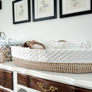 May include: A white and brown wicker bassinet with a white quilted liner and a white blanket. The bassinet is sitting on a white dresser with brown drawers.