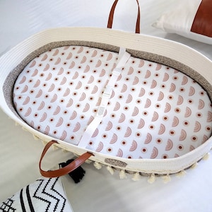 May include: White woven basket with a brown leather handle and a white and pink patterned fitted sheet. The basket has a fringe trim and is perfect for a baby.