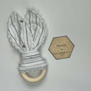 May include: A white and blue striped fabric baby teether with a wooden ring. The fabric has a frayed edge and is tied with a bow. The teether has a small bee design on the fabric. The tag says "Handmade by honey bug love".
