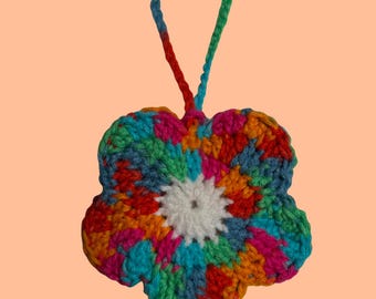 May include: A handmade, colorful crochet flower with a loop for hanging. The flower features a white center and petals in shades of blue, green, orange, pink, and red. The hanging loop transitions from blue to green to red.