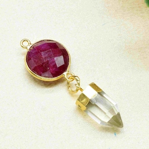 May include: A gold-tone pendant featuring a round, faceted ruby-red gemstone and a clear quartz crystal point.