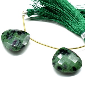 Op de afbeelding: Twee groene edelsteen hangers die met een gouden draad aan elkaar zijn verbonden. De edelstenen zijn gefacetteerd en hebben een donkergroen en zwart gevlekt patroon. De draad is bevestigd aan een groene kwast.