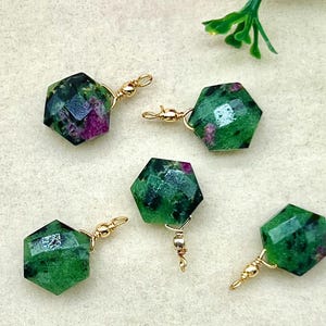 May include: Five faceted, hexagonal earrings with a green and red speckled pattern. Each earring is attached to a gold-coloured hook. The earrings are displayed on a cream-coloured surface, with pink and white flowers in the background.