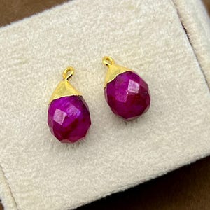 Könnte beinhalten: Zwei facettierte, tropfenförmige Ohrringe in tiefem Magenta. Jeder Ohrring hat eine goldfarbene Kappe. Die Ohrringe werden auf einer cremefarbenen, rechteckigen Schmuckschatulle vor einem braunen Hintergrund präsentiert.