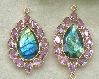 May include: Two gold-toned teardrop-shaped pendants. Each pendant features a central, iridescent, blue-green stone surrounded by smaller, pink, teardrop-shaped stones. The pendants are likely earrings or necklace charms.