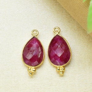 May include: Two gold-tone teardrop-shaped pendants with red gemstone accents. Each pendant has a small gold bead detail at the bottom.
