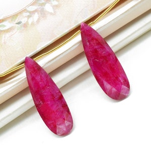 Puede incluir: Dos piedras preciosas de rubí facetadas en forma de lágrima con un tono rosa intenso. Las piedras preciosas están enhebradas en una cadena dorada.