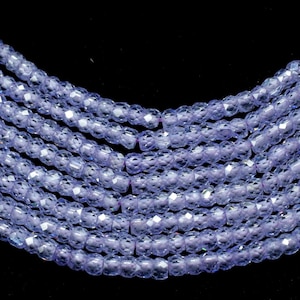 May include: Close-up of several strands of faceted, light purple beads. The beads are small and evenly sized, with a sparkling, reflective surface. The strands are arranged in a gentle curve against a black background, suggesting a necklace or similar jewelry.