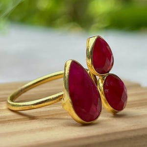 Op de afbeelding: Een goudkleurige ring met drie druppelvormige, gefacetteerde, rode edelstenen. De ring ligt op een houten oppervlak met een vage groene achtergrond.