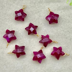 Puede incluir: Primer plano de varias gemas facetadas en forma de estrella, de un tono magenta intenso. Cada estrella está cableada individualmente con alambre dorado, algunas conectadas en cadena. Las gemas están dispuestas sobre una superficie clara.