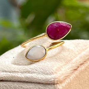 May include: A gold-toned ring featuring two teardrop-shaped gemstones. One gemstone is a deep red, and the other is a pale, milky white. The ring is set against a soft, off-white fabric, with a blurred green background.