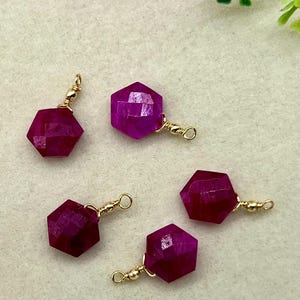 Puede incluir: Cinco colgantes de piedras preciosas magenta oscuro, hexagonales y facetados, con herrajes dorados. Las piedras preciosas son de un color rico y saturado. Los colgantes son probablemente para la fabricación de joyas.