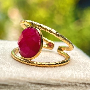 K&ouml;nnte beinhalten: Goldfarbener Ring mit einem gro&szlig;en, oval geschliffenen Rubin. Der Ring hat ein strukturiertes Band und ein einzigartiges, offenes Design. Der Rubin hat eine tiefe, leuchtend rote Farbe. Der Ring liegt auf einer weichen, cremefarbenen Oberfl&auml;che.