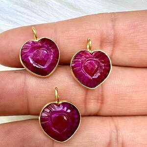 May include: Three heart-shaped pendants with a deep magenta hue and gold-colored trim. Each pendant has a small loop at the top for hanging. The pendants are held in a hand against a white, fluffy background.