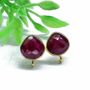 May include: A pair of ruby red gemstone earrings with gold settings. The earrings are shaped like a teardrop and have a small gold loop at the bottom.