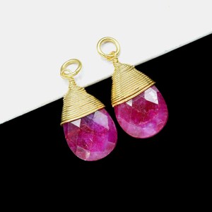 Può includere: Due pietre preziose a forma di goccia di rubino con filo d'oro e un anello d'oro nella parte superiore. I rubini sfaccettati sono di un colore rosso-rosato intenso. Lo sfondo è bianco e nero.