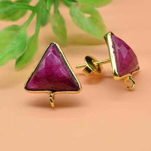 May include: A pair of gold triangle earrings with pink gemstone accents. The earrings have a post back and a small loop on the bottom.