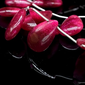 May include: A strand of pink gemstone beads strung on a silver wire. The beads are teardrop shaped and have a glossy finish.