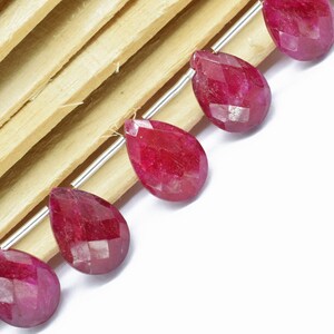 Op de afbeelding: Een streng facetgeslepen robijn edelsteen kralen in een druppelvorm. De kralen zijn geregen op een zilveren draad en worden getoond op een houten oppervlak.