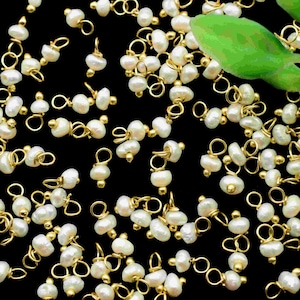 Könnte beinhalten: Eine Sammlung von weißen Perlen- und goldfarbenen Metallperlenanhängern. Jeder Anhänger besteht aus zwei weißen Perlen und einer goldfarbenen Metallschaufel.