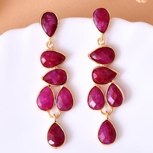 May include: A pair of dangle earrings with five teardrop-shaped ruby red gemstones on each earring. The earrings are made of gold-toned metal.