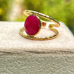 K&ouml;nnte beinhalten: Ein goldfarbener Ring mit einem gro&szlig;en, oval geformten, tiefroten Edelstein. Der Ring hat ein strukturiertes Band und ein Doppelband-Design. Der Edelstein ist in einer goldenen L&uuml;nette gefasst. Der Ring wird auf einer hellen Oberfl&auml;che pr&auml;sentiert.