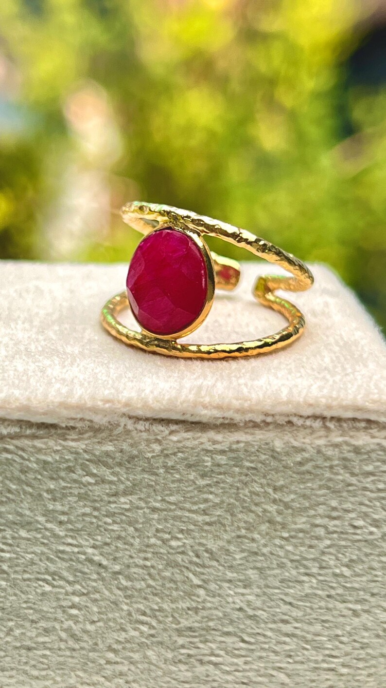 K&ouml;nnte beinhalten: Ein goldfarbener Ring mit einem gro&szlig;en, oval geformten, tiefroten Edelstein. Der Ring hat ein strukturiertes Band und ein Doppelband-Design. Der Edelstein ist in einer goldenen L&uuml;nette gefasst. Der Ring wird auf einer hellen Oberfl&auml;che pr&auml;sentiert.