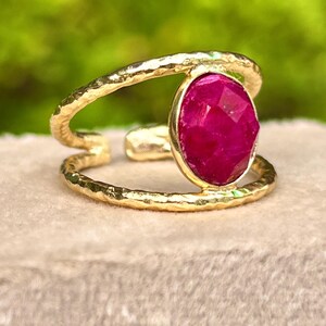 K&ouml;nnte beinhalten: Ein goldfarbener Ring mit einem gro&szlig;en, facettierten, ovalen, magentafarbenen Edelstein. Der Ring hat ein Doppelband-Design mit gehammerter Textur. Der Edelstein ist in einer goldenen L&uuml;nette gefasst. Der Ring wird auf einer neutralfarbenen Oberfl&auml;che pr&auml;sentiert.