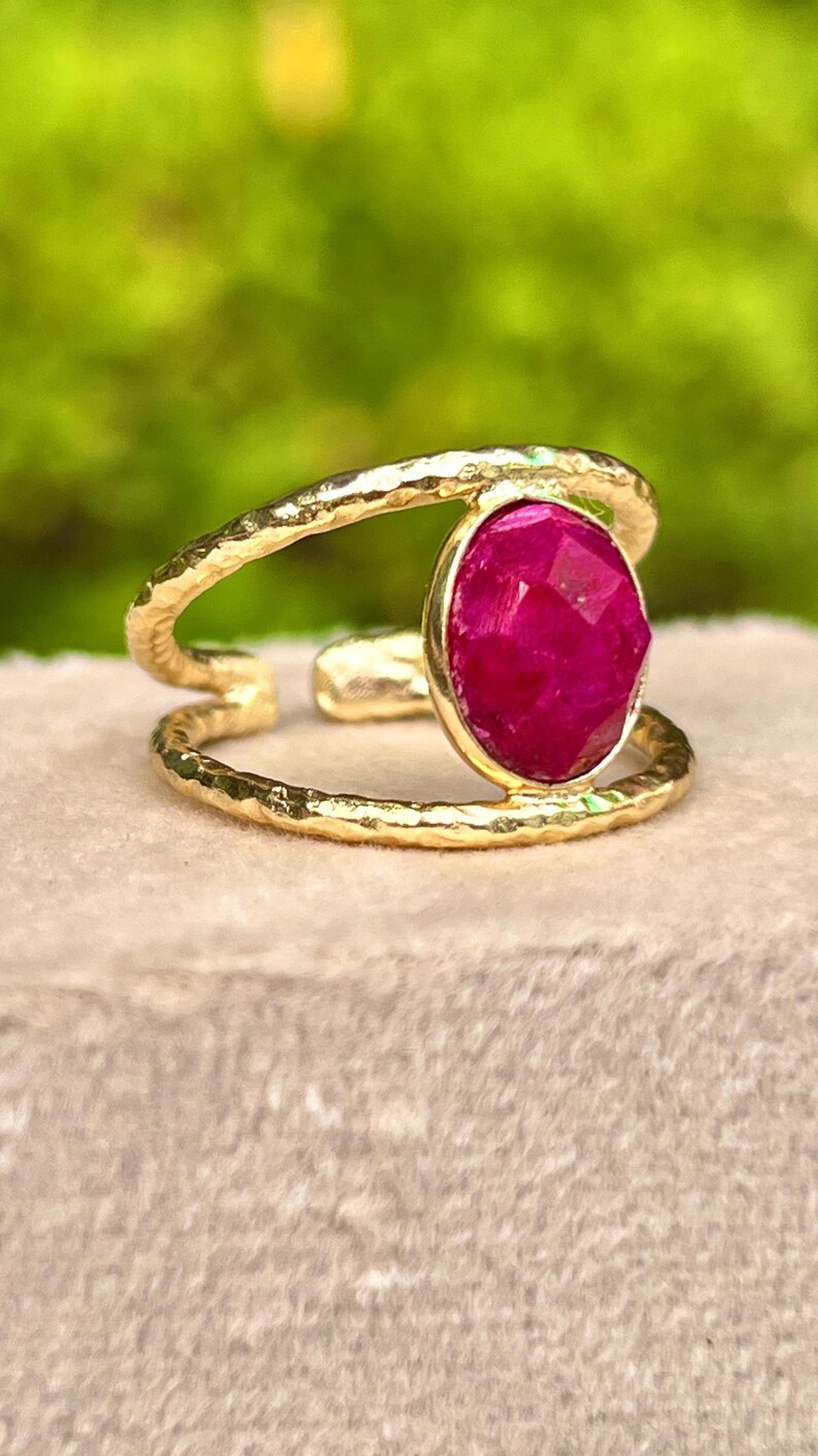 K&ouml;nnte beinhalten: Ein goldfarbener Ring mit einem gro&szlig;en, facettierten, ovalen, magentafarbenen Edelstein. Der Ring hat ein Doppelband-Design mit gehammerter Textur. Der Edelstein ist in einer goldenen L&uuml;nette gefasst. Der Ring wird auf einer neutralfarbenen Oberfl&auml;che pr&auml;sentiert.