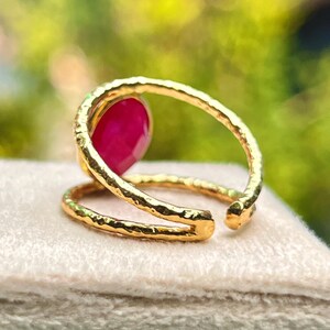 K&ouml;nnte beinhalten: Ein goldfarbener Ring mit einem gro&szlig;en, oval geformten, roten Edelstein. Der Ring hat ein strukturiertes Band, das sich um den Finger wickelt. Der Ring wird auf einer hellen Oberfl&auml;che mit verschwommenem gr&uuml;nem Hintergrund pr&auml;sentiert.