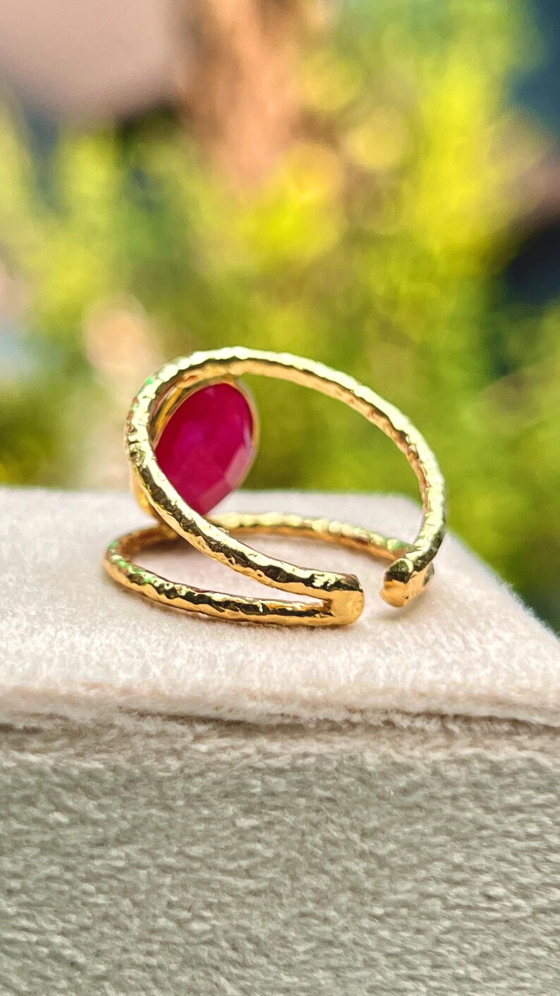 K&ouml;nnte beinhalten: Ein goldfarbener Ring mit einem gro&szlig;en, oval geformten, roten Edelstein. Der Ring hat ein strukturiertes Band, das sich um den Finger wickelt. Der Ring wird auf einer hellen Oberfl&auml;che mit verschwommenem gr&uuml;nem Hintergrund pr&auml;sentiert.