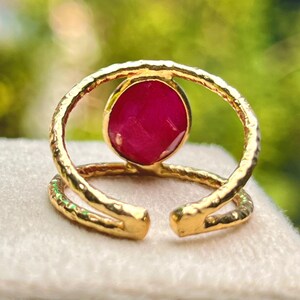 K&ouml;nnte beinhalten: Ein goldfarbener Ring mit einem gro&szlig;en, runden, tiefroten Edelstein in der Mitte. Der Ring hat ein strukturiertes Band und ein offenes Design. Der Edelstein ist in einer L&uuml;nettenfassung gefasst. Der Ring wird auf einer hellen Oberfl&auml;che pr&auml;sentiert.