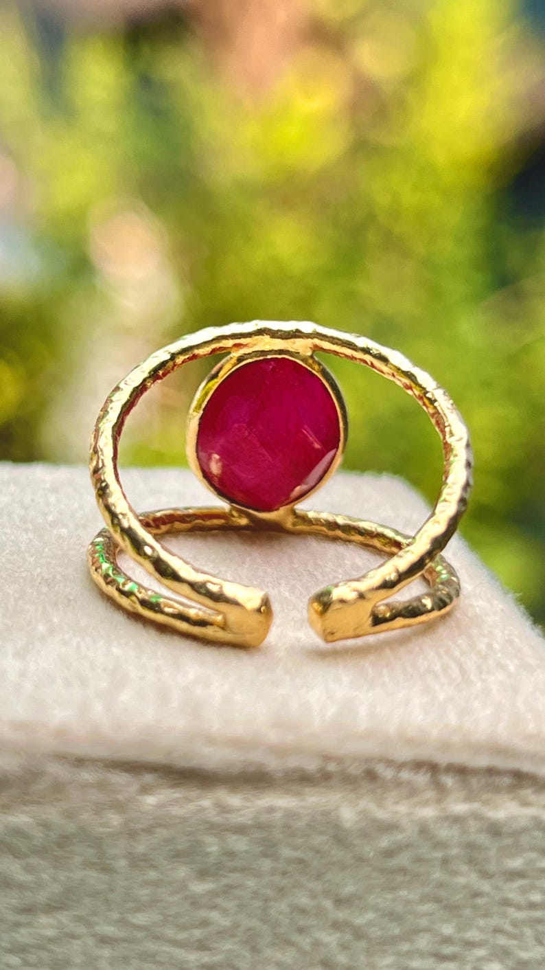 K&ouml;nnte beinhalten: Ein goldfarbener Ring mit einem gro&szlig;en, runden, tiefroten Edelstein in der Mitte. Der Ring hat ein strukturiertes Band und ein offenes Design. Der Edelstein ist in einer L&uuml;nettenfassung gefasst. Der Ring wird auf einer hellen Oberfl&auml;che pr&auml;sentiert.