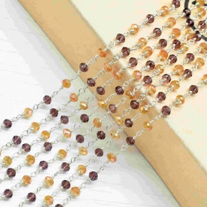 Pode incluir: Uma corrente de prata com contas facetadas alternadas de roxo e laranja. A corrente está disposta sobre uma superfície marrom.