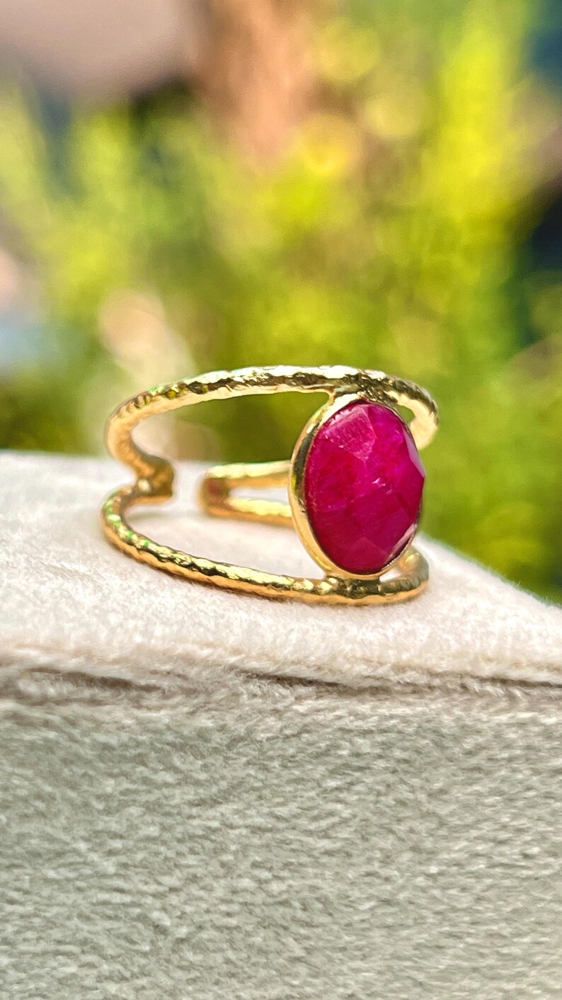 K&ouml;nnte beinhalten: Ein goldfarbener Ring mit einem gro&szlig;en, oval geformten, facettierten Rubin-Edelstein. Der Ring hat ein strukturiertes Band und ein geteiltes Schaftdesign. Der Rubin hat einen leuchtenden Rotton. Der Ring wird auf einer hellen Oberfl&auml;che pr&auml;sentiert.