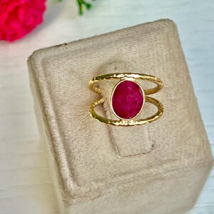 K&ouml;nnte beinhalten: Goldfarbener Ring mit einem gro&szlig;en, oval geschliffenen Rubin. Der Ring hat ein strukturiertes Band und ein Doppelband-Design, wobei der Rubin in der Mitte gefasst ist. Der Ring wird auf einer beigen Oberfl&auml;che mit Blumen im Hintergrund pr&auml;sentiert.