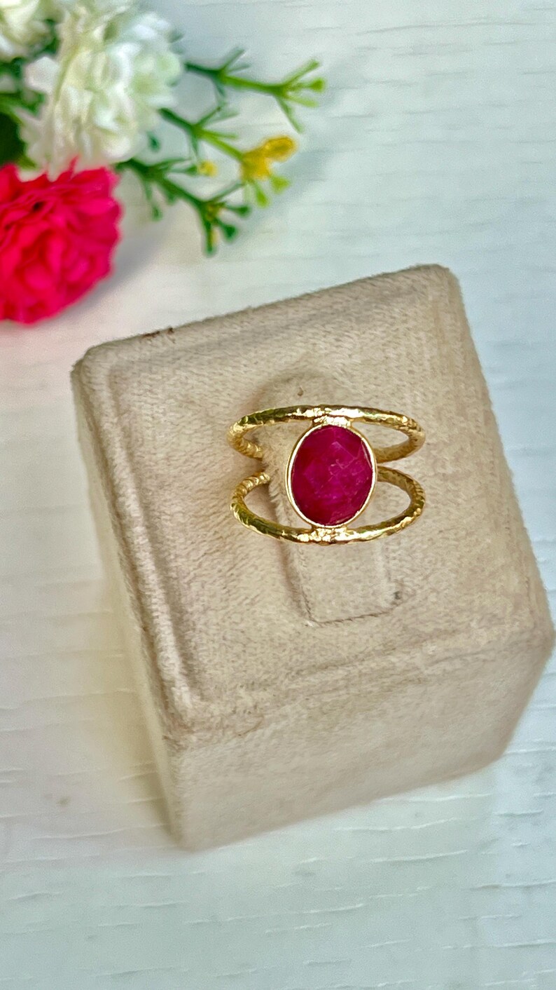 K&ouml;nnte beinhalten: Goldfarbener Ring mit einem gro&szlig;en, oval geschliffenen Rubin. Der Ring hat ein strukturiertes Band und ein Doppelband-Design, wobei der Rubin in der Mitte gefasst ist. Der Ring wird auf einer beigen Oberfl&auml;che mit Blumen im Hintergrund pr&auml;sentiert.