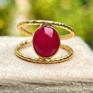 K&ouml;nnte beinhalten: Ein goldfarbener Ring mit einem gro&szlig;en, ovalen, tiefroten Edelstein. Der Ring hat ein Doppelband-Design mit strukturierter Oberfl&auml;che. Der Edelstein ist der Mittelpunkt, gefasst in einer goldenen L&uuml;nette.