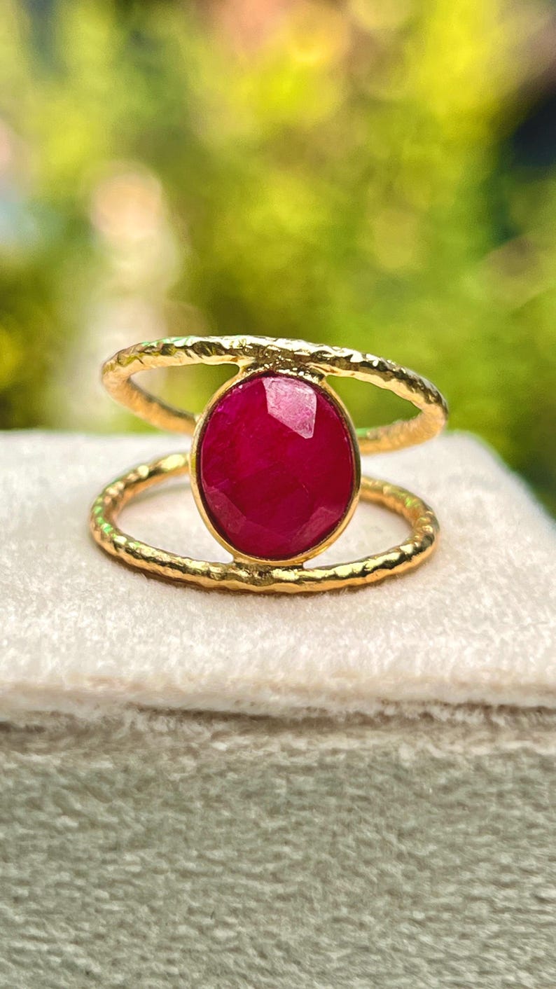 K&ouml;nnte beinhalten: Ein goldfarbener Ring mit einem gro&szlig;en, ovalen, tiefroten Edelstein. Der Ring hat ein Doppelband-Design mit strukturierter Oberfl&auml;che. Der Edelstein ist der Mittelpunkt, gefasst in einer goldenen L&uuml;nette.