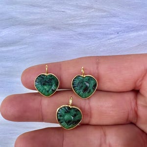 May include: Three heart-shaped pendants with a carved design and a vibrant green hue. Each pendant is encased in a gold-coloured setting with a small loop at the top for hanging. The pendants are displayed against a soft, white background.