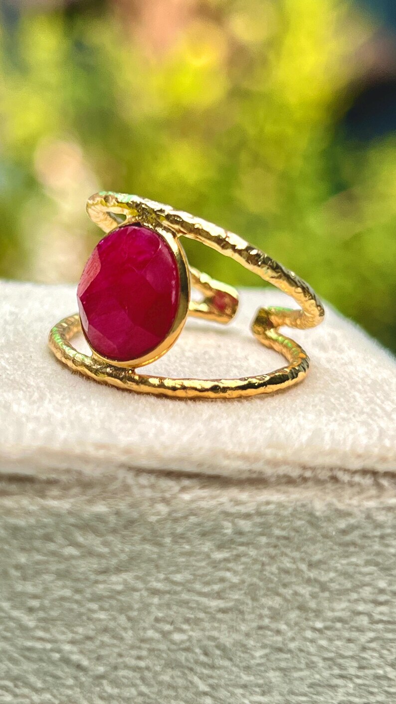K&ouml;nnte beinhalten: Goldfarbener Ring mit einem gro&szlig;en, oval geschliffenen Rubin. Der Ring hat ein strukturiertes Band und ein einzigartiges, offenes Design. Der Rubin hat eine tiefe, leuchtend rote Farbe. Der Ring liegt auf einer weichen, cremefarbenen Oberfl&auml;che.